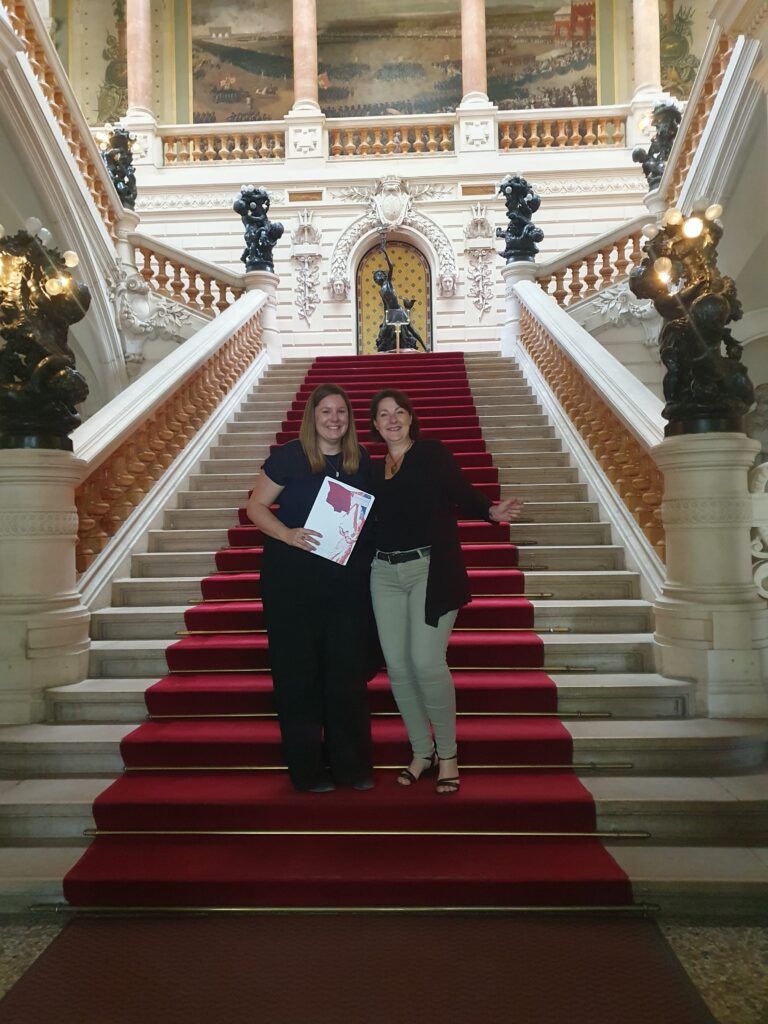 Two women pose on a grand marble staircase with a red carpet and ornate statues flanking the steps.